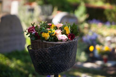 Korbtasche mit bunten Rosen steht auf einem Grab, im Hintergrund sind unscharfe Blumen und Grabsteine sichtbar.