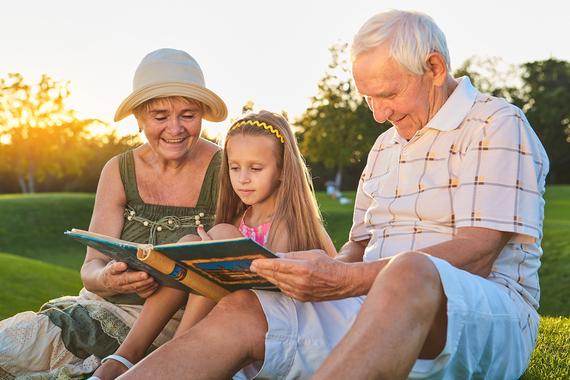 Großeltern und Enkelin sitzen im Freien und lesen gemeinsam in einem Buch bei Sonnenuntergang.